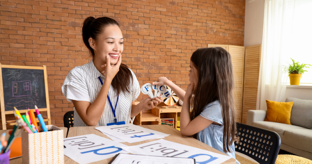 A teacher and a young girl are sitting at a table engaging in a language learning activity focused on sh words for speech therapy. The teacher smiles as she assists the girl, who holds up letters. The room is bright and decorated with a brick wall and a small blackboard.